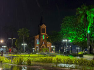 #PraTodosVerem: imagem da Igreja Matriz de Santa Isabel Rainha de Portugal, localizada no Centro do Paulista, durante a noite. A cena mostra a igreja iluminada enquanto a chuva cai sobre a região, com o céu escuro e reflexos das luzes na via molhada ao redor do templo.