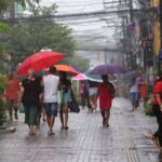 #PraTodosVerem: pessoas caminham pela Rua Siqueira Campos durante a chuva, utilizando sombrinhas e guarda-chuvas para se proteger. A via aparece molhada, com reflexos da iluminação nas poças d’água e movimentação de pedestres ao longo da rua.