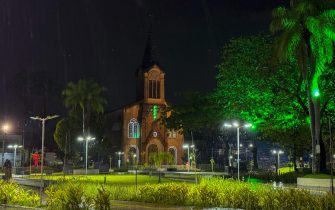 #PraTodosVerem: imagem da Igreja Matriz de Santa Isabel Rainha de Portugal, localizada no Centro do Paulista, durante a noite. A cena mostra a igreja iluminada enquanto a chuva cai sobre a região, com o céu escuro e reflexos das luzes na via molhada ao redor do templo.