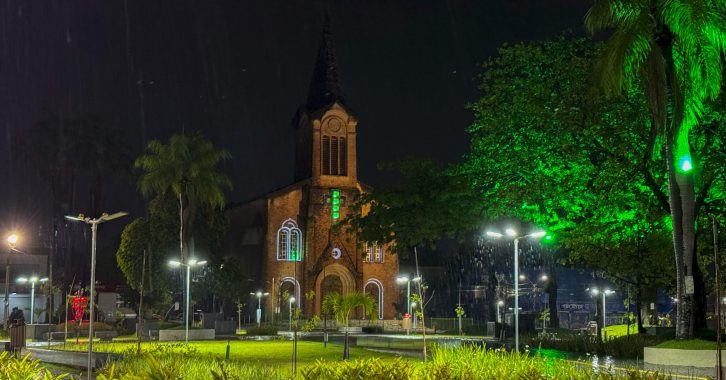 #PraTodosVerem: imagem da Igreja Matriz de Santa Isabel Rainha de Portugal, localizada no Centro do Paulista, durante a noite. A cena mostra a igreja iluminada enquanto a chuva cai sobre a região, com o céu escuro e reflexos das luzes na via molhada ao redor do templo.